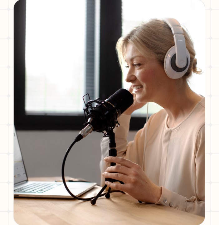 A professional woman wearing headphones, smiling and speaking into a podcast microphone at a sunlit desk.